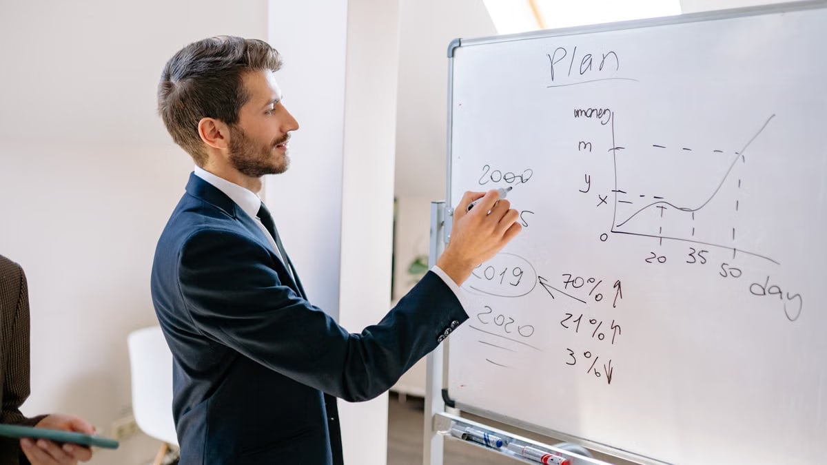 A businessman in a suit writes financial data on a whiteboard during an office planning session.